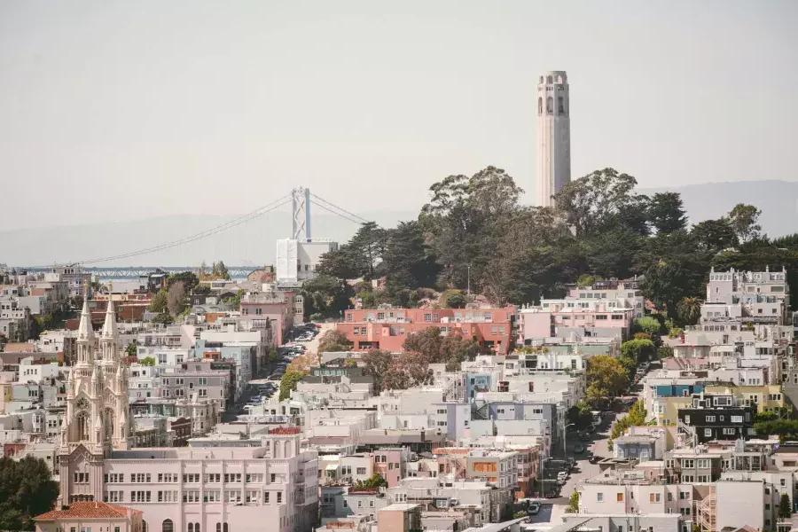 A Coit Tower de São Francisco é retratada com a Bay Bridge ao fundo e uma colina coberta de casas em primeiro plano.