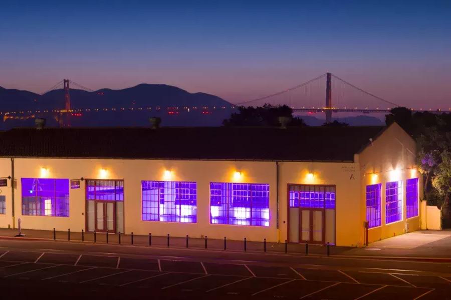 Das Fort Mason-Gebäude wird nachts mit einer violetten Innenbeleuchtung beleuchtet, im Hintergrund ist die Brücke zu sehen.